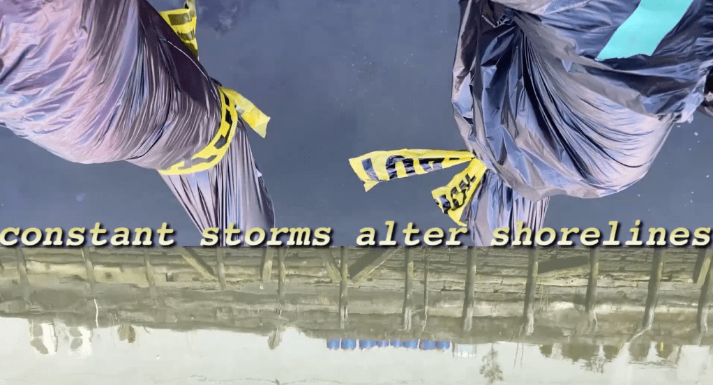 Still from video speculating the future of the Gowanus Canal shorreline. Includes reflectiion in the water and feet encased in black garbage bags dangling, with caution tape, and the words "constant storms alter shorelines" superimposed in yellow serif font